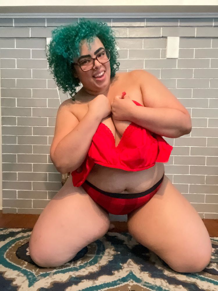 Cruel Valentine, a Black, non-binary person with teal hair and eyebrows, tortoise shell glasses, and a nose ring, gives the camera an open-mouthed grin as they kneel, legs apart, wearing red and black hunter’s plaid panties and clutching a newly removed red bralette to cover their chest, exposing just a titillating amount of cleavage.