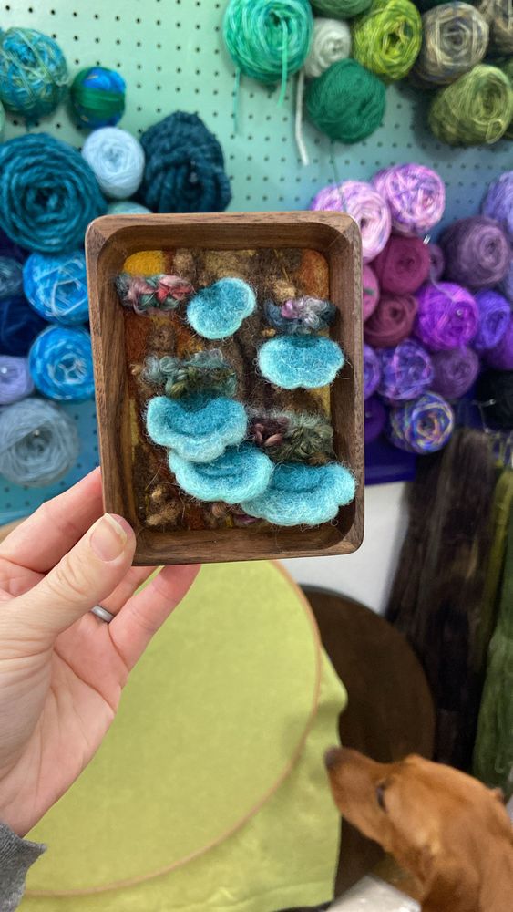 A left hand holds up a small, rectangular piece of art in a brown, wooden frame. The art depicts Forest floor scenery with blue, bracket mushrooms. In the background there is a colorful wall of yarn and an out of focus dog sniffing the wall. 