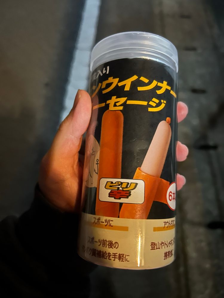 Hand holding a plastic tube with an illustration of Popeye the sailor on it next to a sausage. In this one one of the sausages is like half unskinned for some reason. If you can read Japanese it says it’s spicy