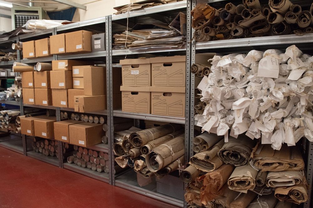 A view of archives on a shelving unit. There are various boxes, as well as documents rolled up and stacked in groups.
