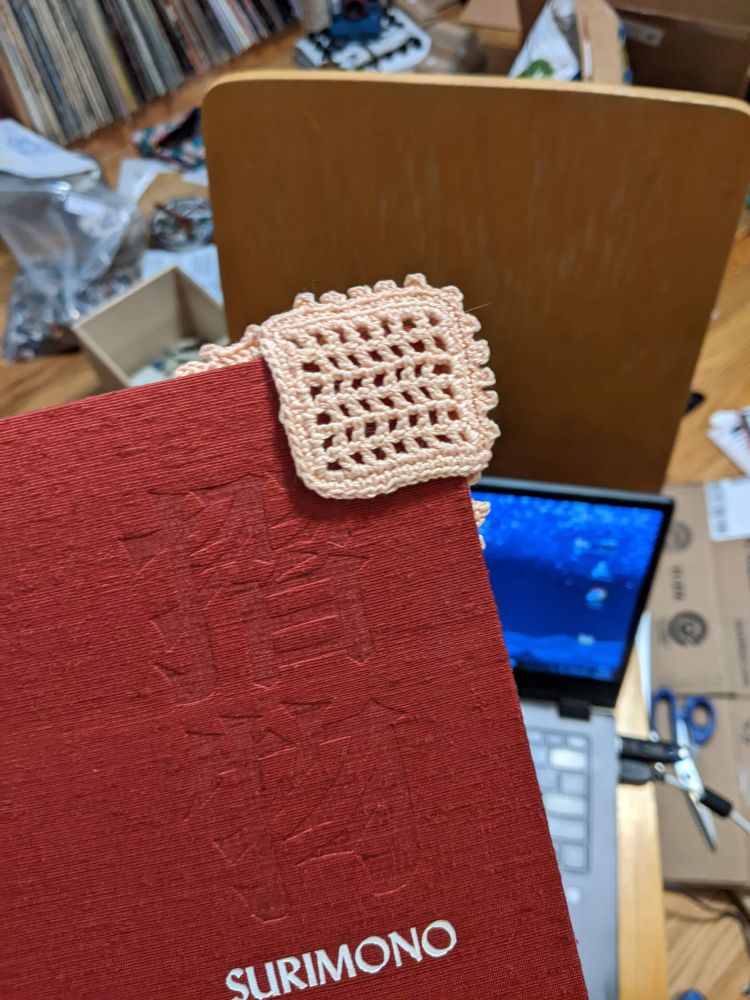 Image shows a red book cover with a bookmark on the top corner. The bookmark is lace like knit work and is the shape of a square on this side! It's also a dusty pink and very beautiful! 