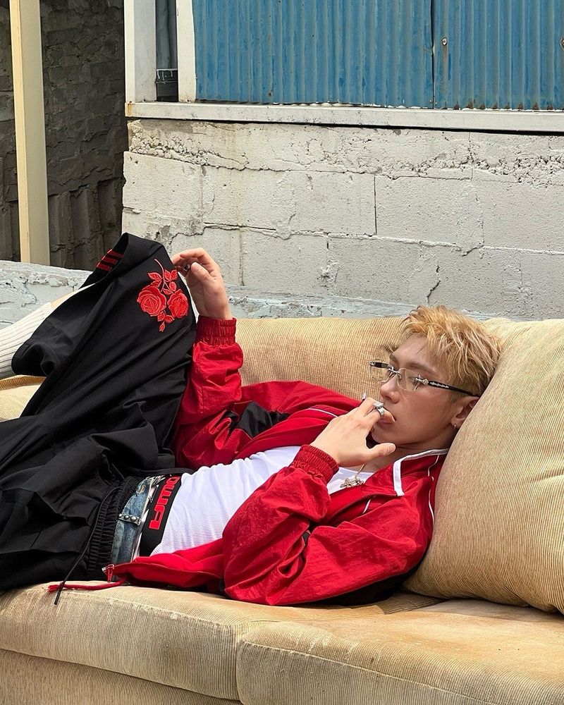 WayV Ten laying down on a yellow couch with one leg over the armrest. He’s wearing a glasses, a red jacked over a white tank top and black over the knee shorts. He’s holding his left hand to his mouth and the right one hovering over his knee.