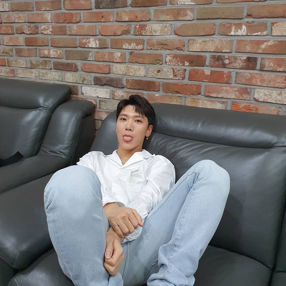 WAYV TEN LEE sitting on a black couch, looking beautiful wearing a white button down and light blue jeans, sticking his tongue out playfully 