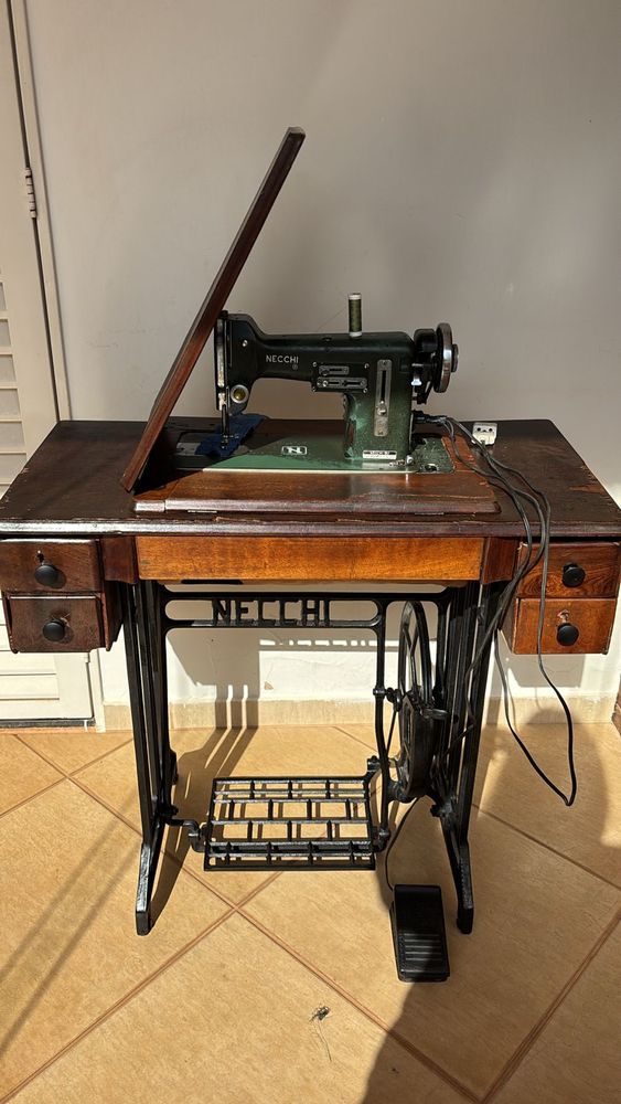 Máquina de costura antiga Necchi (verde-oliva) embutida numa mesa de madeira, com base de ferro fundido e pedal de balanço onde se lê “NECCHI”.