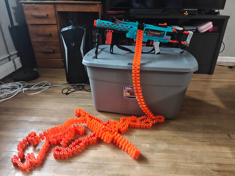 Photo of a dart zone boxer pro, a foam dart blaster made to resemble a belt fed machine gun. The blaster is on top of a box, and it has a very long belt of ammunition attached to it. The belt is mostly piled up on the floor. 
