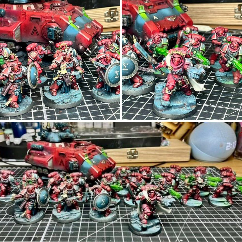 A professionally painted Warhammer 40,000 Primaris Space Marines army is displayed on a hobbyist’s cutting mat, showcasing exceptional miniature painting skill. The force features Primaris Marines in striking crimson red armor with contrasting white and teal highlights, creating a bold and cohesive color scheme. At the front, a squad of elite warriors with ornate shields and detailed bases stand ready for battle, flanked by Marines wielding green-glowing plasma and bolt weaponry. A heavily customized red Primaris Repulsor tank with teal accents and turret-mounted weaponry looms in the background, adding armored support to the force. The scene is set in a well-equipped hobby workspace with visible paint bottles, brushes, and tools in the background. This image highlights the craftsmanship, detail, and artistry of Warhammer 40k miniature painting, ideal for showcasing painted Space Marines, Primaris army displays, and hobby setups
