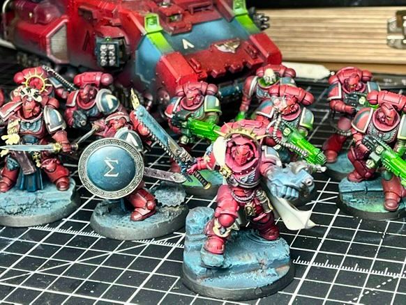 A professionally painted Warhammer 40,000 Primaris Space Marines army is displayed on a hobbyist’s cutting mat, showcasing exceptional miniature painting skill. The force features Primaris Marines in striking crimson red armor with contrasting white and teal highlights, creating a bold and cohesive color scheme. At the front, a squad of elite warriors with ornate shields and detailed bases stand ready for battle, flanked by Marines wielding green-glowing plasma and bolt weaponry. A heavily customized red Primaris Repulsor tank with teal accents and turret-mounted weaponry looms in the background, adding armored support to the force. The scene is set in a well-equipped hobby workspace with visible paint bottles, brushes, and tools in the background. This image highlights the craftsmanship, detail, and artistry of Warhammer 40k miniature painting, ideal for showcasing painted Space Marines, Primaris army displays, and hobby setups