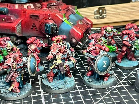 A professionally painted Warhammer 40,000 Primaris Space Marines army is displayed on a hobbyist’s cutting mat, showcasing exceptional miniature painting skill. The force features Primaris Marines in striking crimson red armor with contrasting white and teal highlights, creating a bold and cohesive color scheme. At the front, a squad of elite warriors with ornate shields and detailed bases stand ready for battle, flanked by Marines wielding green-glowing plasma and bolt weaponry. A heavily customized red Primaris Repulsor tank with teal accents and turret-mounted weaponry looms in the background, adding armored support to the force. The scene is set in a well-equipped hobby workspace with visible paint bottles, brushes, and tools in the background. This image highlights the craftsmanship, detail, and artistry of Warhammer 40k miniature painting, ideal for showcasing painted Space Marines, Primaris army displays, and hobby setups