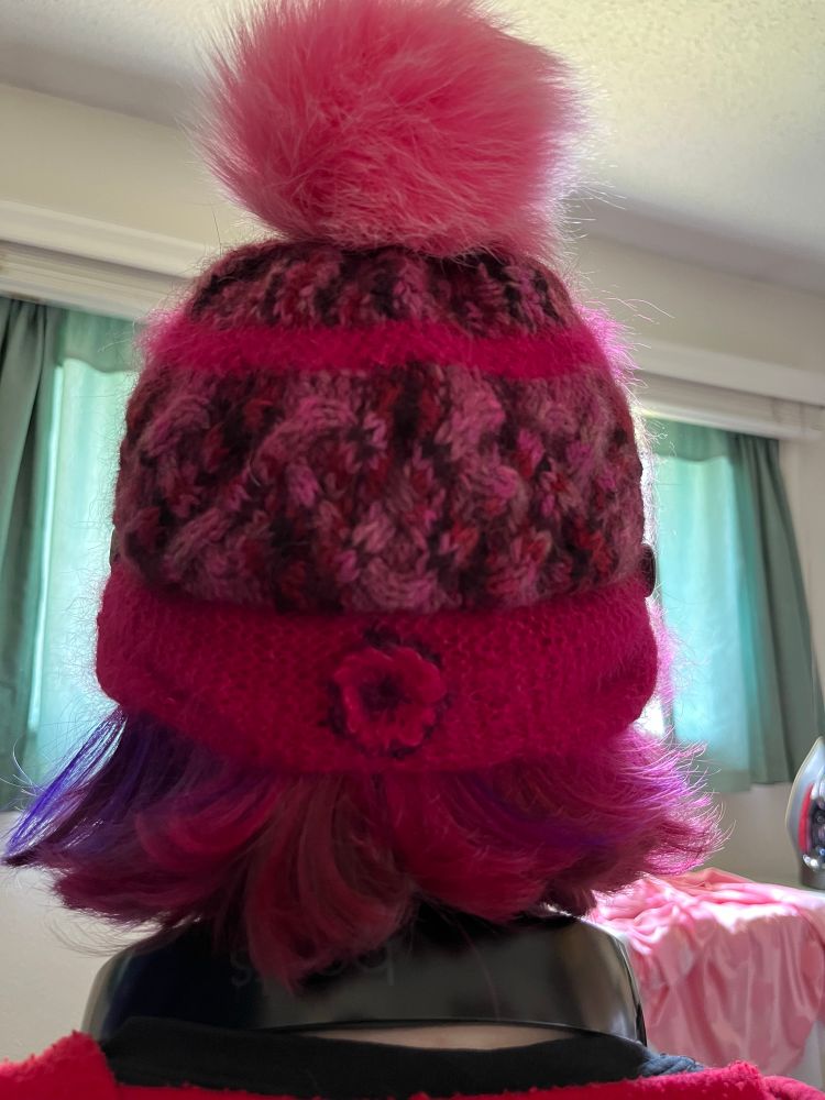 Pink cabled hat with earflaps and a pink pompom, back view eith a crocheted flower detail. 
