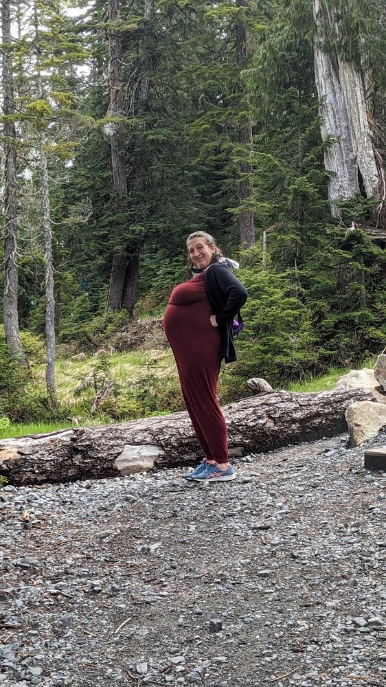 Laura in profile, with her hands on her hips, showing off her pregnant belly, in a forest.