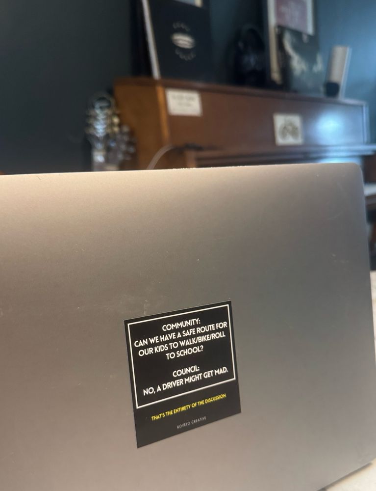 Square sticker on the front of a laptop that says community can we have a safe route for our kids to walk bike and roll to school? Council no a driver Mike get mad that’s the entirety of the discussion.