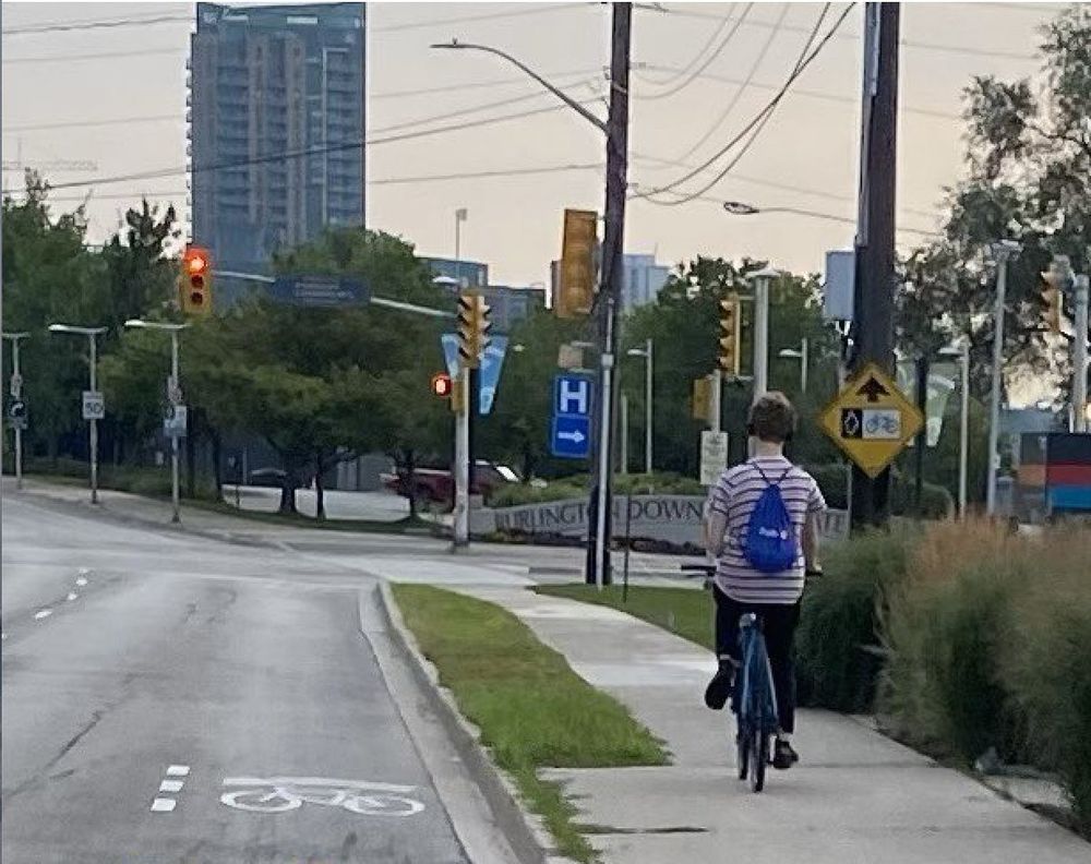 Person riding a bicycle on an empty sidewalk next to a road with three dotted lines and a sharrow. 