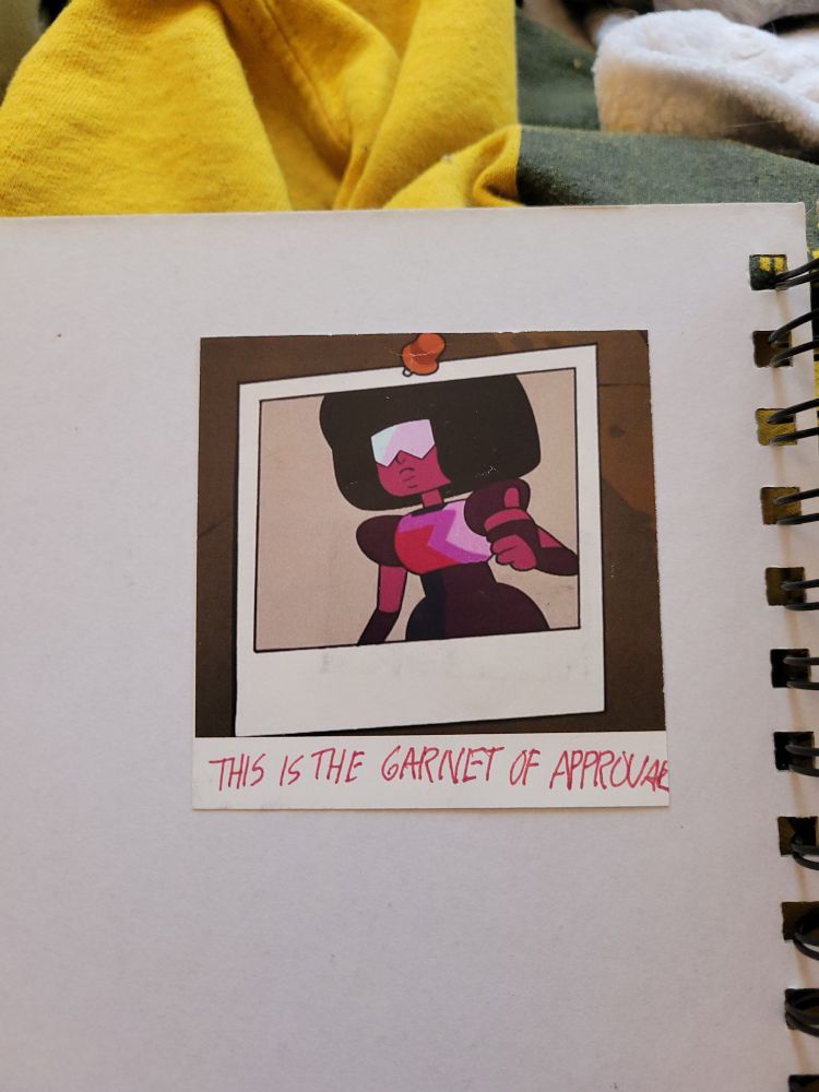 printed out Polaroid of garnet from Steven universe doing a thumbs up. the handwritten note under it states "THIS IS THE GARNET OF APPROVAL" and she guarded the entrance of my senior painting studio until I graduated and now she lives in this sketchbook