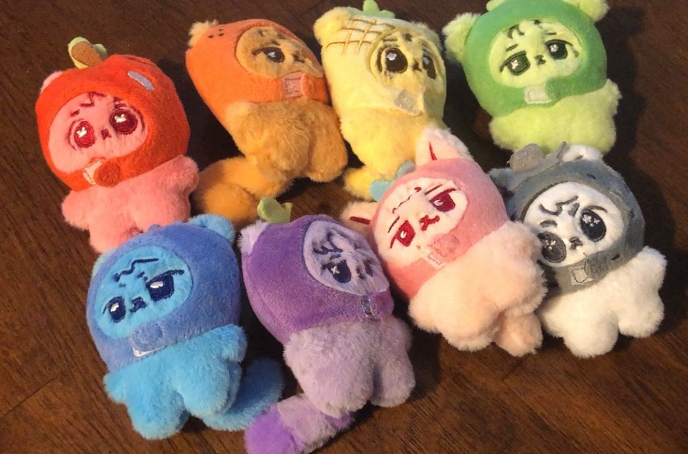 A set of 8 different mini plushies with chubby limbs, main focal color, animal theme, and fruit hat accessory are laid together against a wood background.