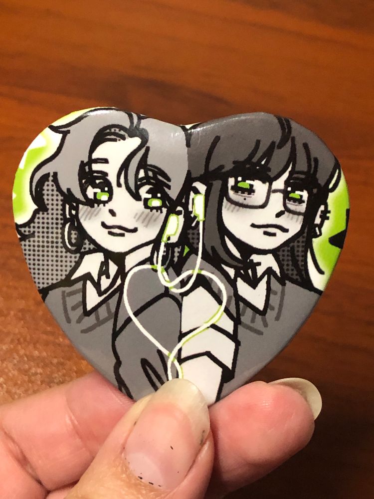 heart shaped green button of aya and mitsuki from "the guy she was interested in wasn't a guy at all." they are shoulder-to-shoulder, sharing a set of earphones and the wires loop into a heart towards the bottom of the design.