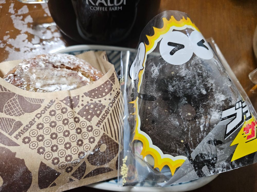 ミスタードーナツのドーナツ二個の写真。紙袋に入った穴の相手ないブリュレドーナツと、顔の書いたビニール袋に入ったブラックサンダーチョコドーナツ。