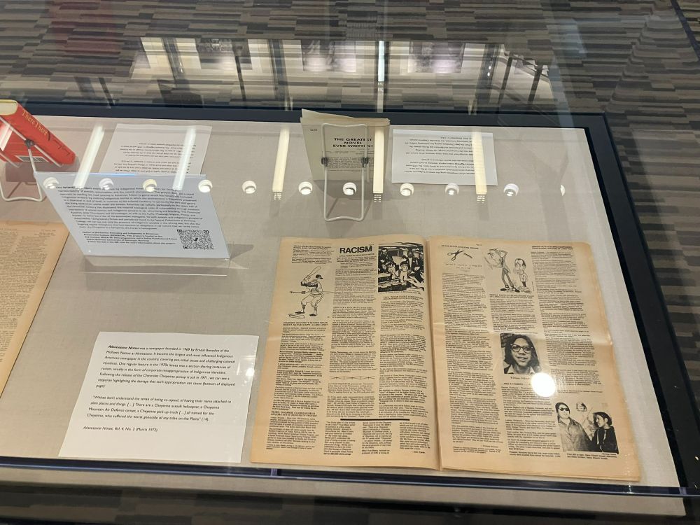 A glass display case containing 1970s Indigenous newspaper Akwesasne Notes, opened to a page with the heading "Racism" and depicting stereotypical media images of Native Americans