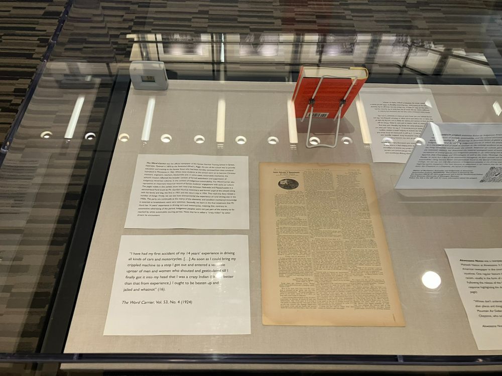 A glass display case containing historical periodical The Word Carrier opened to a page with a photograph of an Indigenous man driving a Ford Truck from 1921