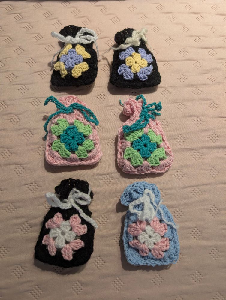 Granny square lavender pouches in different colour combos, from top to bottom, left to right: enby flag colours with purple at the center, then yellow than black, enby flag colours with yellow at the center than purple, 2 identical ones with deep green, light green then pink, one with white, pink then black and one trans flag colours with white at the center, pink then blue 