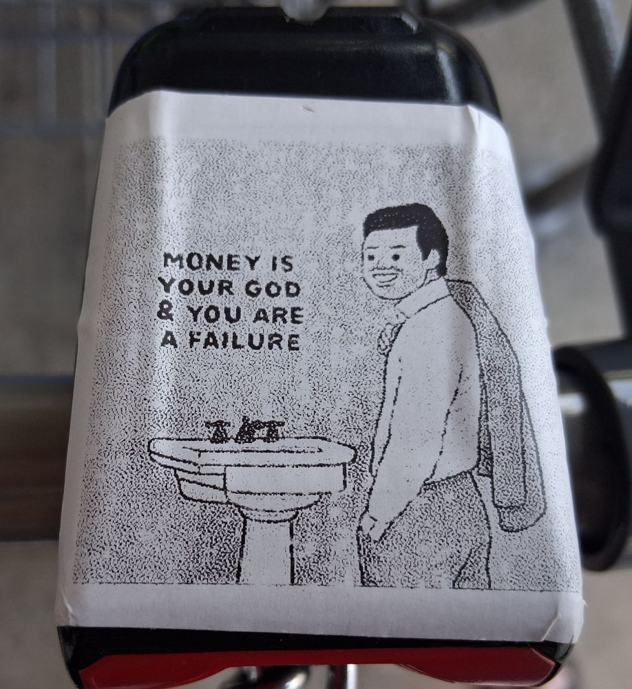 Black and white sticker on top of a coin slot of an Aldi's shopping cart shows a sink and a man in a suit with the jacket over his right shoulder. Text: Money is your god and you are a failure 
