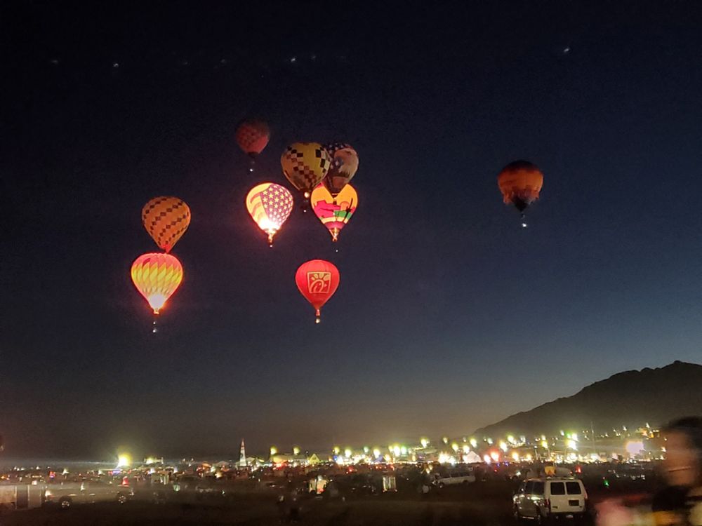 Photo of nine hot air balloons hanging low in the dark pre-dawn sky. A number of them are lit up because the gas is firing. In the lower right-hand side the edge of the Sandia mountains can be seen