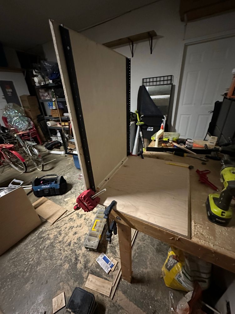 A picture of a wooden cabinet with rack hardware clamped and glued on a workbench.  The garage is messy with a lot of sawdust on the floor and random things in the background.