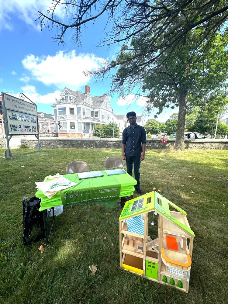 Clean Water Action's intern stands behind a green table and doll house with energy efficiency info on the town green on a sunny day