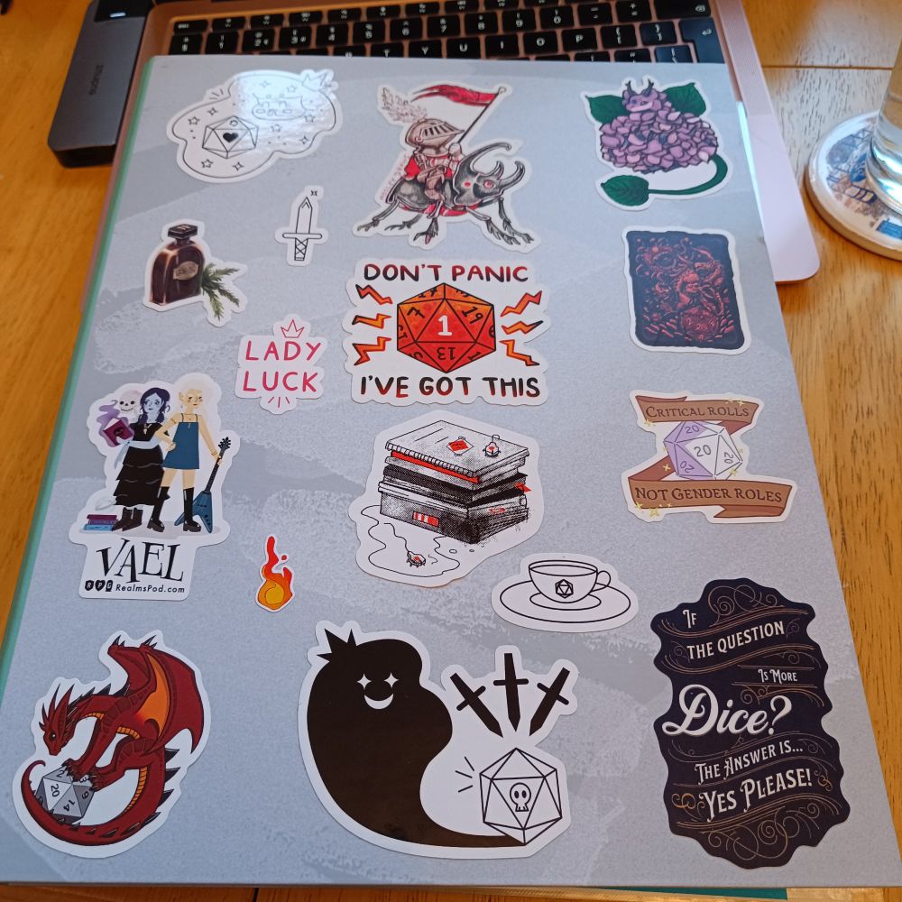 A photo of a binder on a desk covered in TTRPG stickers, including one depicting Isabel and Maerydyth from the podcast Realm's Vael campaign, and a few of Dungeon Delves' depictions of Lady Luck