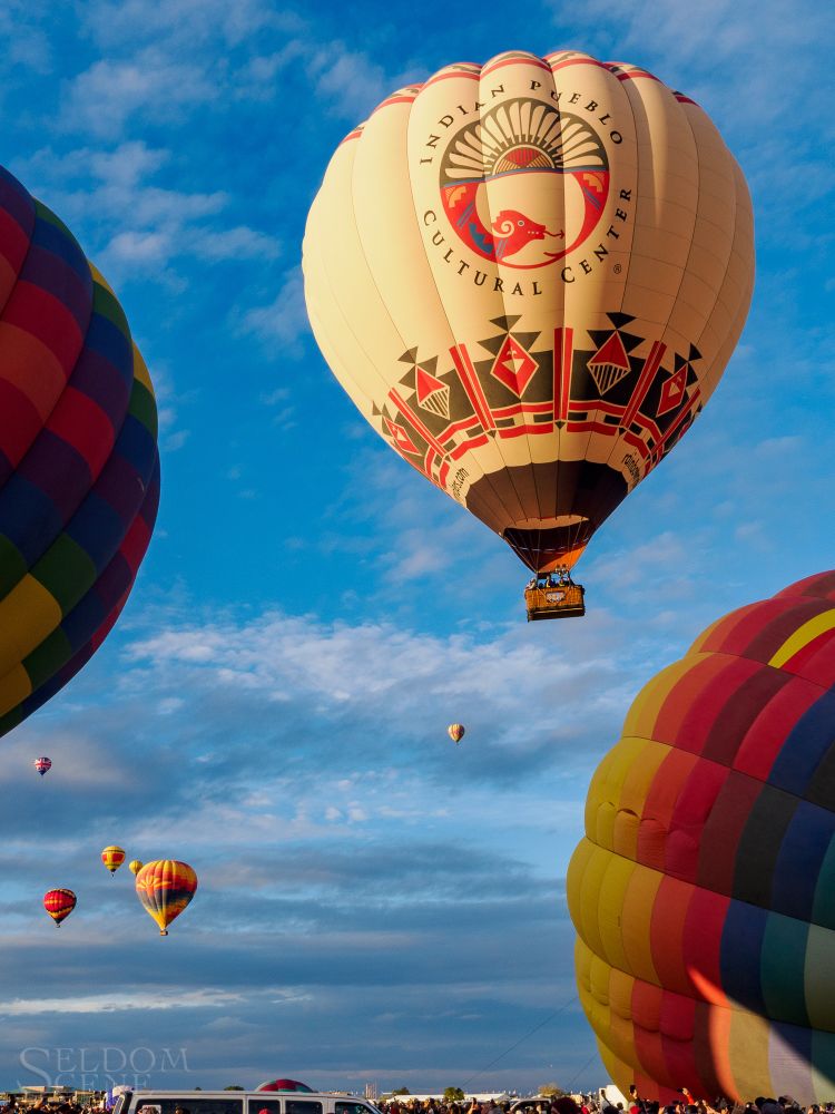 A tan hot air balloon decorated with Pueblo tribal motifs rises into a crisp morning sky, rapidly distancing itself from balloons still on the field.