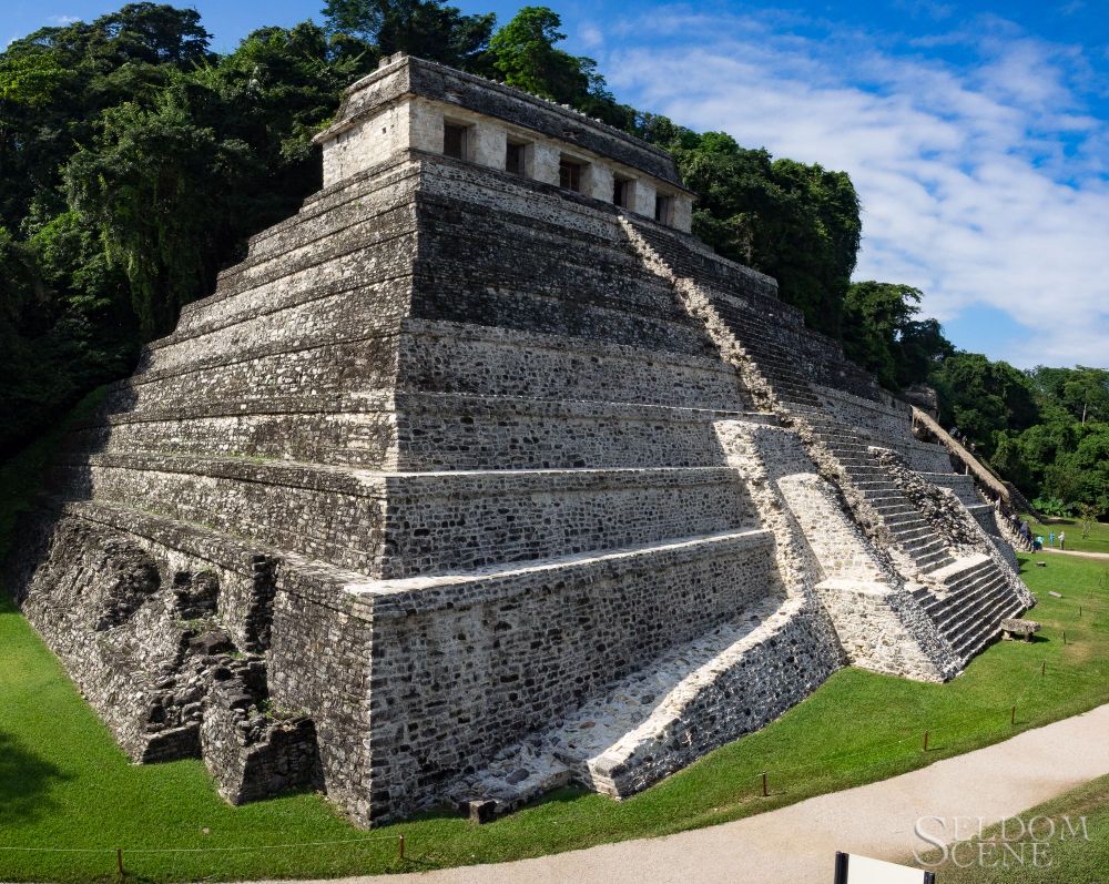 A stepped Maya pyramid shines in the morning sunlight. It's surrounded by green turf, backed by healthy foliage, and overseen by a blue sky with thin clouds.