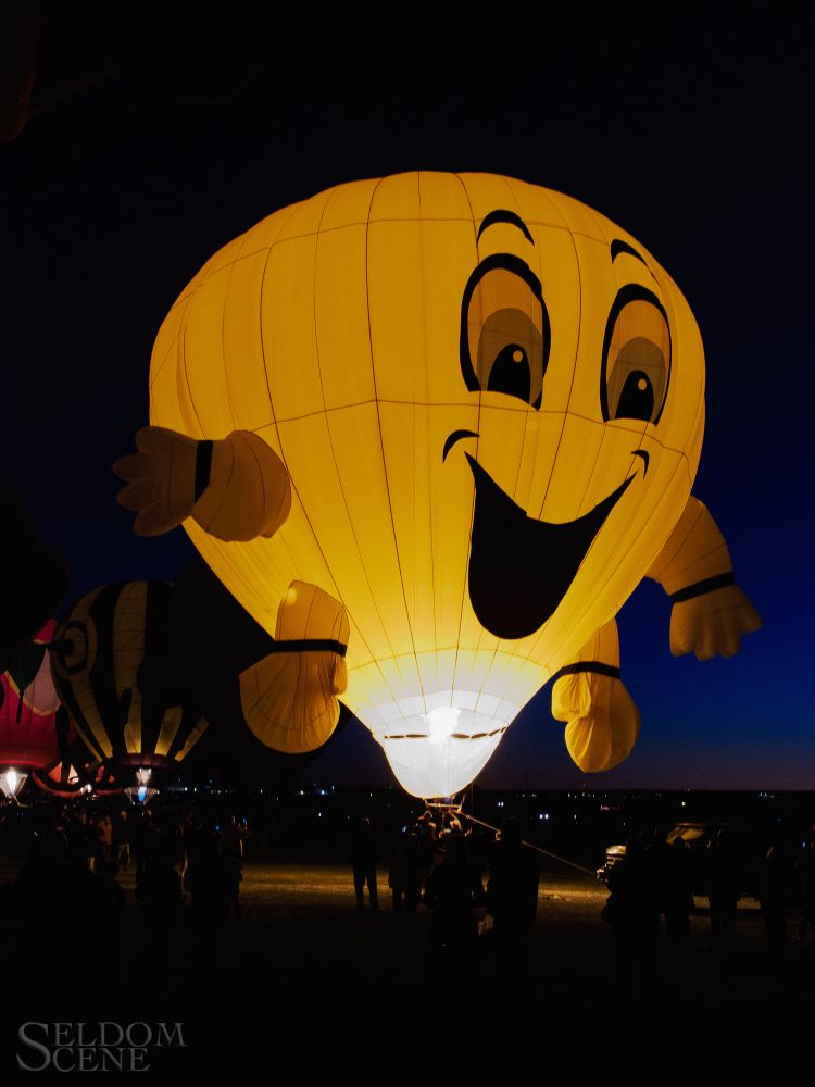 A bright yellow hot air balloon in the form of an enthusiastic, smiling face lights up the night when its burner is ignited.