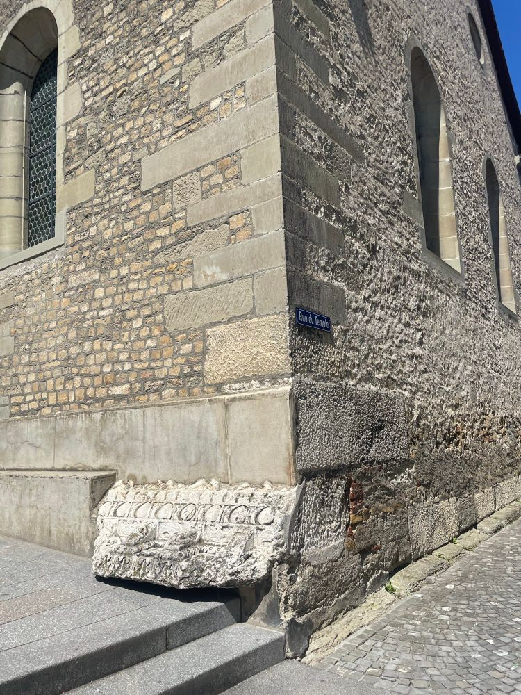 View of the exterior corner of the church of Ste-Marie-Madeleine in Avenches. Most of the church is built of greyish brown stone, and the exterior is plain. But at the bottom corner an extravagant piece of carved Roman masonry in white limestone sticks out, with coffered cornices and egg-and-dart motifs on the modillion. 