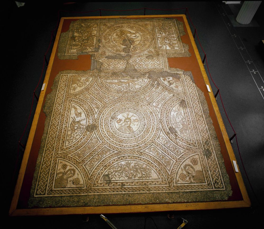 Picture of the full Hinton St Mary mosaic, showing two rectangular panels with figural scenes joined by a narrow geometric panel. The central roundel of the larger panel shows a male figure with a chi-rho behind him; the corners contain images of the Four Seasons. The central roundel of the smaller panel shows Bellerophon fighting the Chimaera. Hunting scenes flank both roundels. 