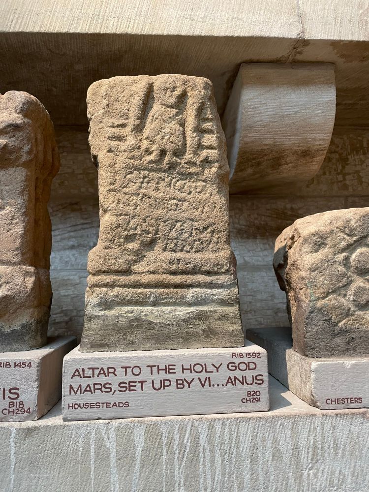 Small Roman altar, with a relief of a roughly carved cloaked male figure standing in a niche. Below is a worn inscription. Both the figure and the inscription are noticeably at a slant. The modern museum base reads “Altar to the holy god Mars, set up by Vi…anus”, RIB 1592 from Housesteads. The altar sits on a shelf, with other carved stones visible to either side. 