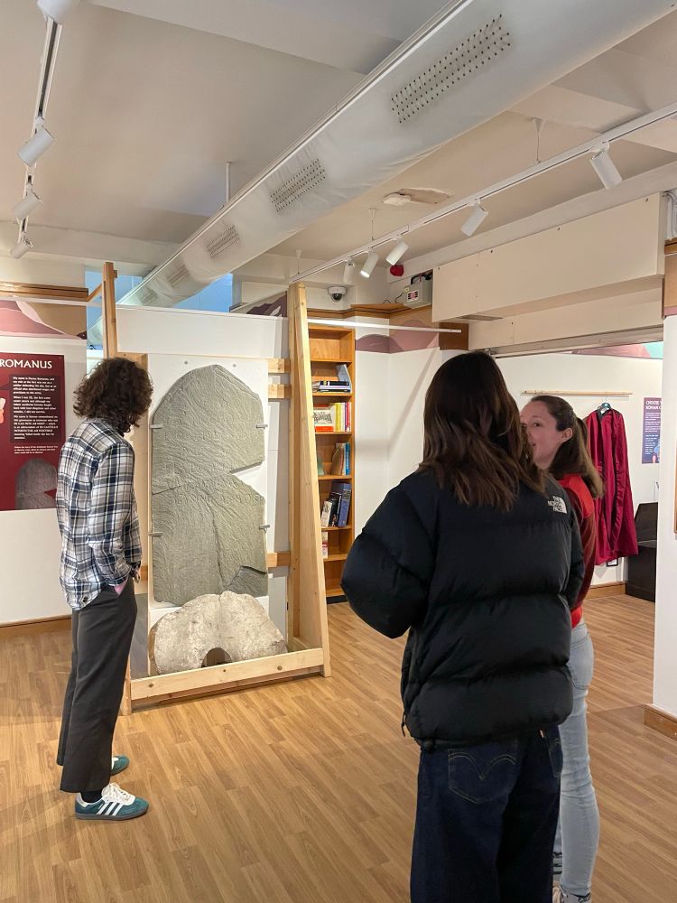Rowan Colley, Isla Mustin and Faye Morissey discussing the tombstone of Flavius Fuscinus and Flavius Romanus, held in The Armitt 