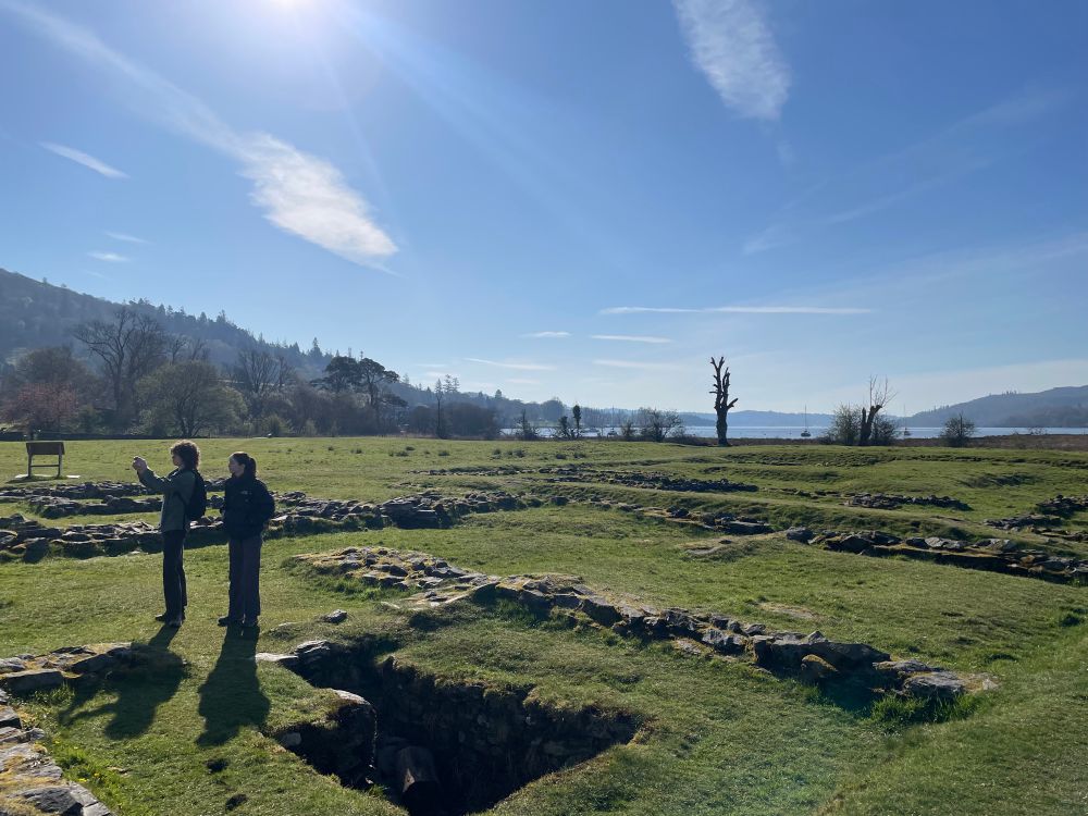 View of the principia at Ambleside, under a bright blue sky with Windermere in the background. Two students stand in the principia taking pictures. 