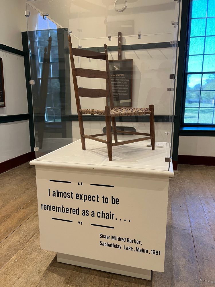Photo of a shaker chair in a display case. The pedestal of the case has the words i almost expect to be remembered as a chair, a quote by a shaker sister in maine. 