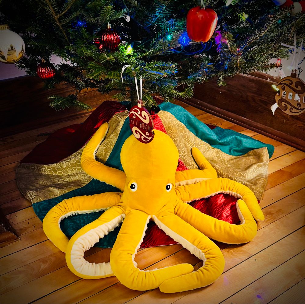 A plush yellow octopus, under a Christmas tree.