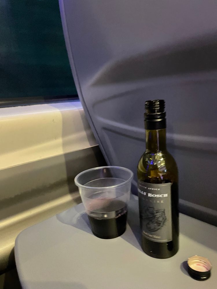 Small bottle of red wine next to a plastic glass of same next to a train window 