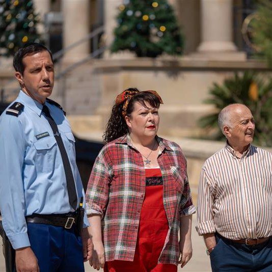 Caron, Gloria and Uncle Patrick stand in front of a building with Christmas trees. Caron is in his Gendarme uniform, Gloria wears a plaid shirt over a red dress, and Uncle Patrick wears a striped shirt. They all looking attentively in the same direction.
