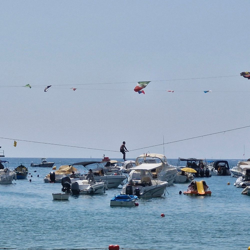 A man in swimming shorts straddles a rope strung across a bay. There are small boats and people in the water.