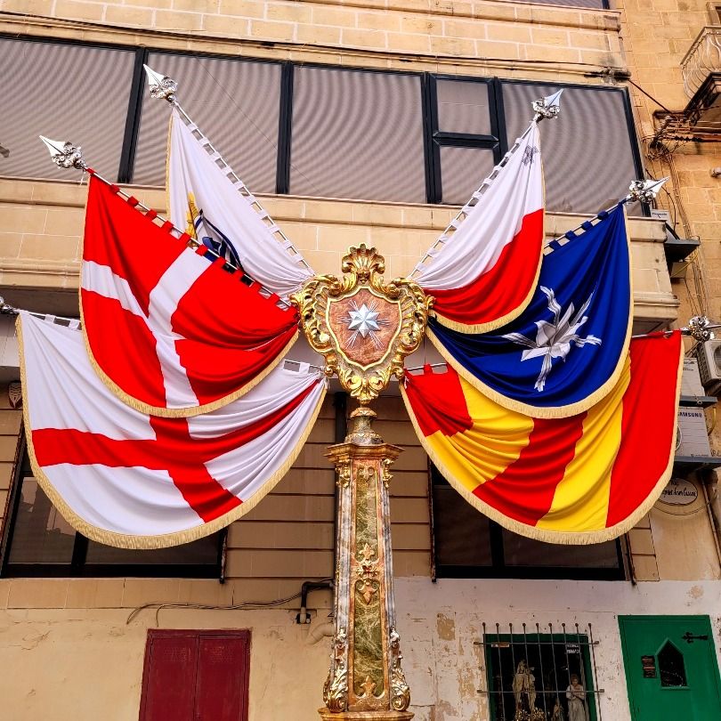 A decorative pole adorned with six large, colourful flags featuring the flags of St George, the Knights of St John and Malta, set against a stone building.