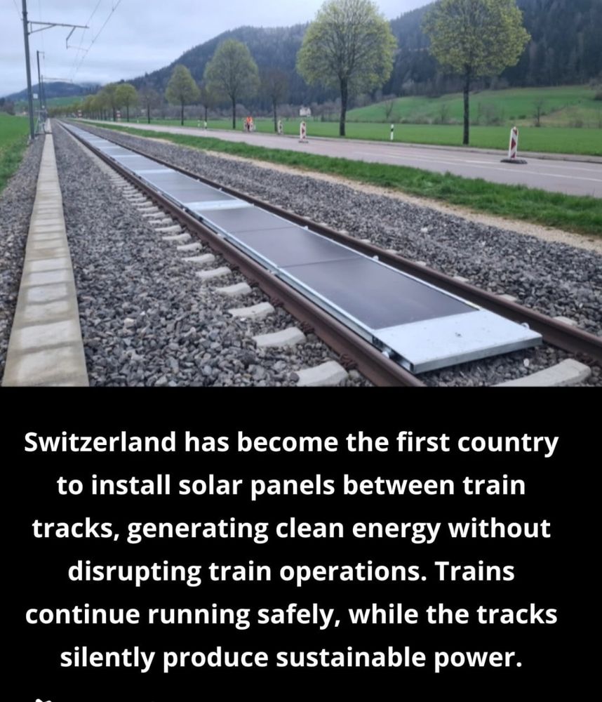 A photo shows a railway track in Switzerland where solar panels have been installed between the rails. The panels are flat and aligned with the track, allowing trains to pass over them. The setting is rural, with green fields, a road, and rows of trees alongside the track, backed by forested hills under a cloudy sky. Text below the image states that Switzerland is the first country to install solar panels between train tracks, enabling clean energy generation without disrupting train operations.