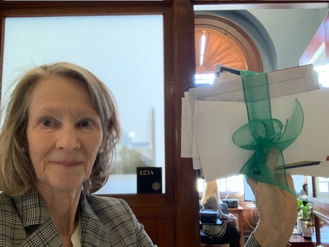 Photo of Kate McGovern (an older white woman) outside RI Governor Dan McKee's office holding up a handful of cards tied together with a green bow.