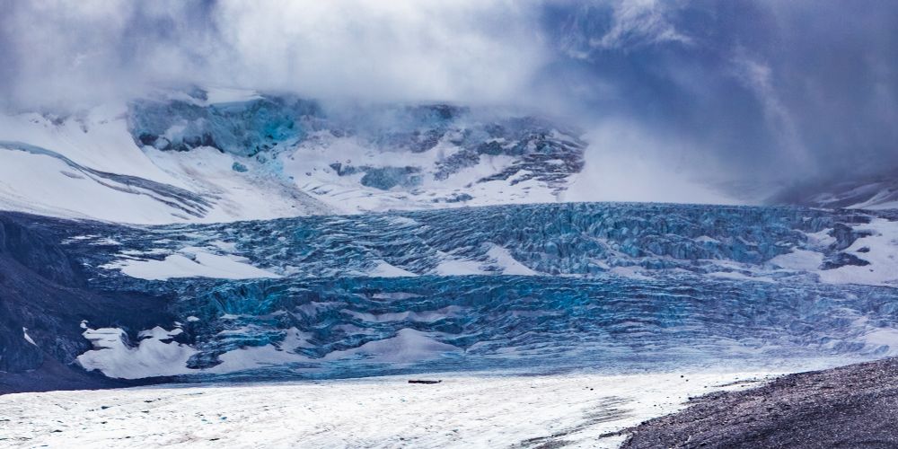 A dramatic view of a glacier with layers of blue ice and snow cascading down a mountain slope under a cloudy sky. The scene is cold and rugged, showcasing the natural power and beauty of glacial landscapes.
