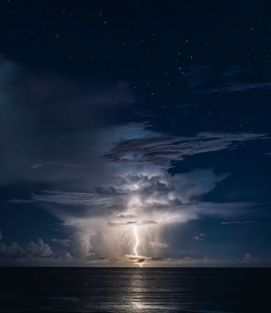 A powerful lightning bolt strikes the ocean, illuminating a dramatic thundercloud at night. The sky is filled with stars, and the lightning creates a bright reflection on the calm, dark water below. The cloud structure is layered and towering, adding to the intensity of the scene.