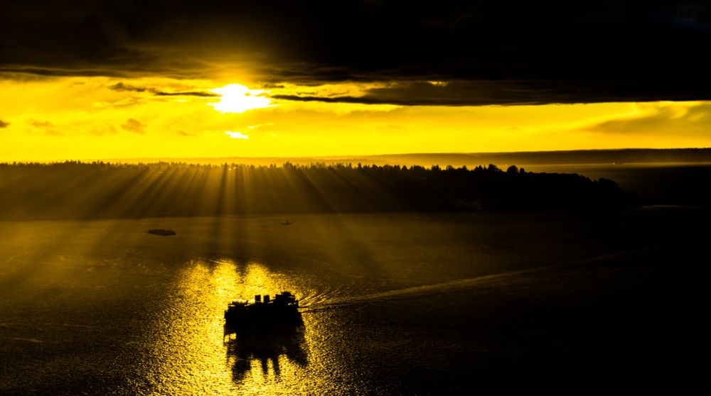 A ferry sails across shimmering waters under dramatic golden rays of sunlight breaking through dark clouds at sunset, casting long reflections and creating a striking silhouette against the glowing horizon.