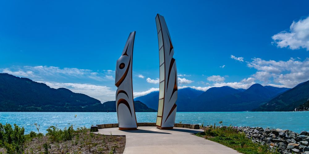 Two tall, stylized Indigenous art sculptures stand on a waterfront pathway with a turquoise lake and mountain range in the background under a bright blue sky.