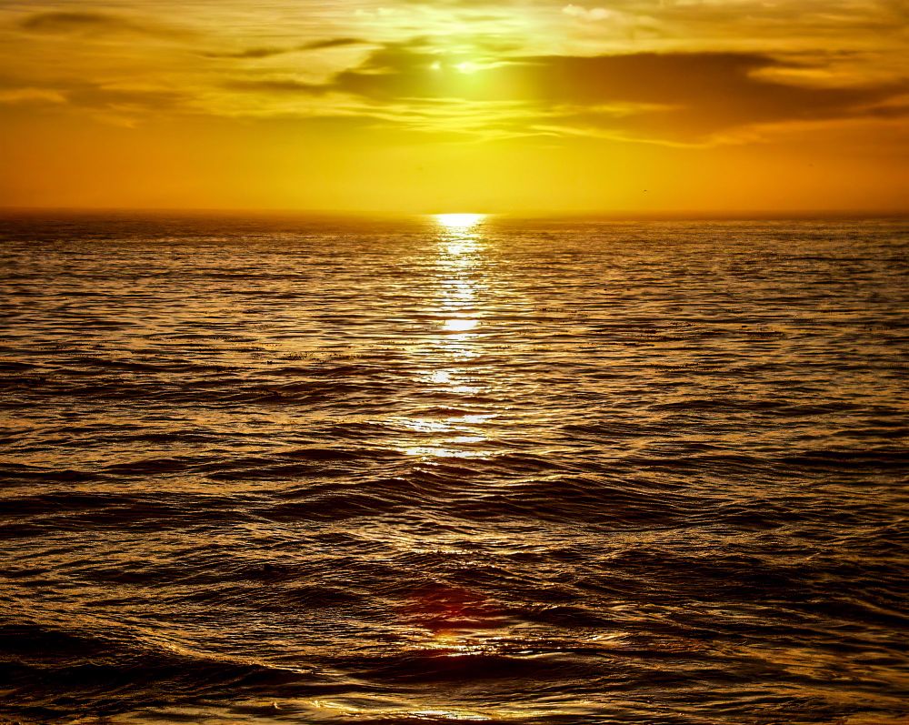 A vivid golden sunset over a gently rippling ocean, with the sun low on the horizon casting a bright reflection across the water and illuminating the sky with rich orange and yellow tones.
