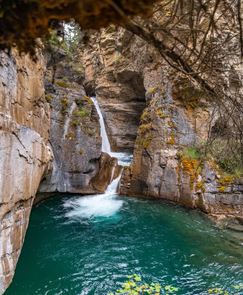 A narrow waterfall cascades through rugged rock cliffs into a swirling turquoise pool, surrounded by steep canyon walls.